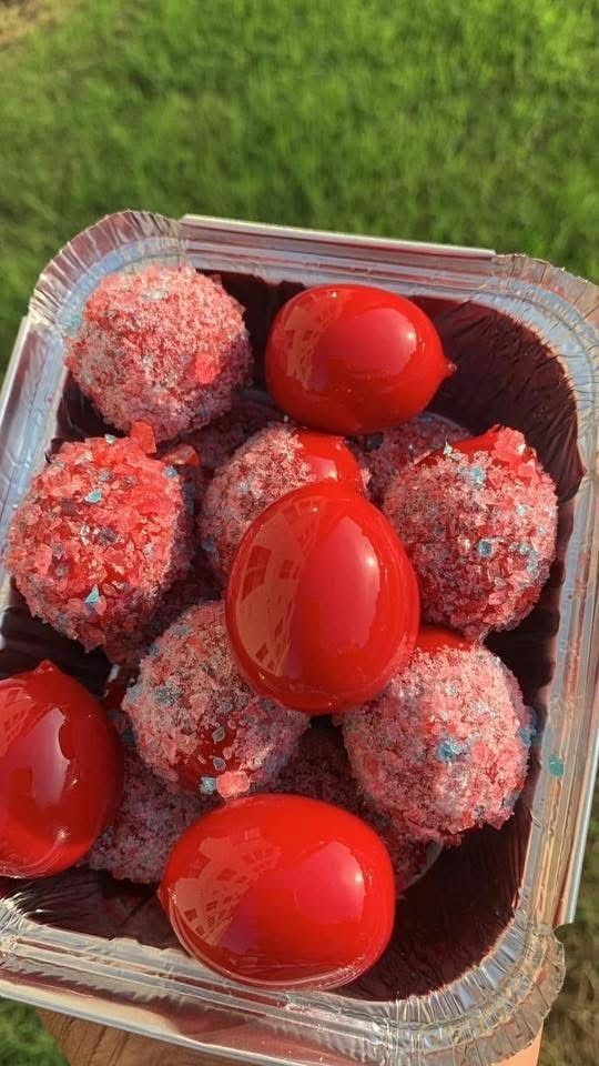 A container holds a mix of red, shiny glazed treats and sugar-coated, textured round sweets against a grassy background.