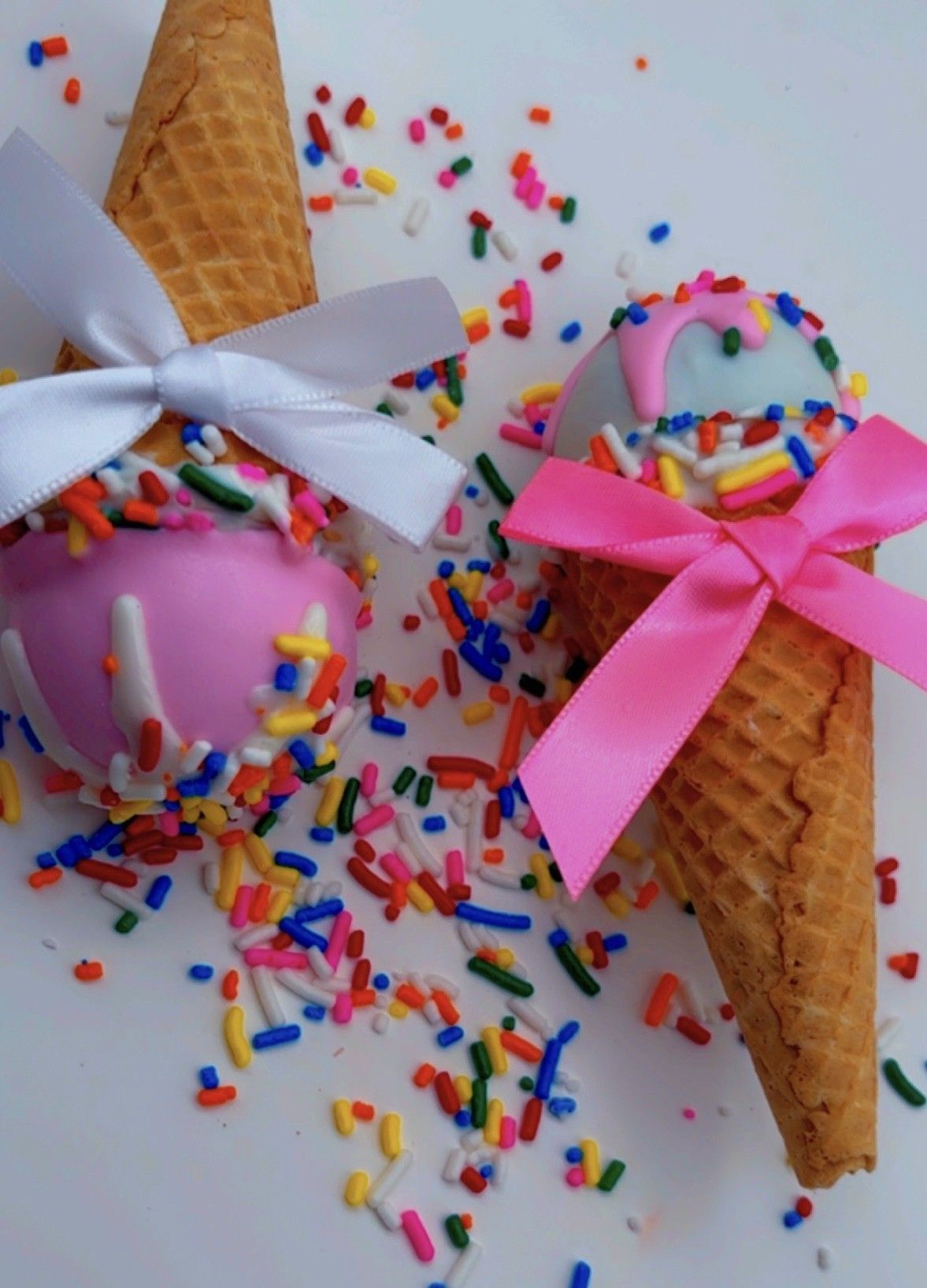 Two ice cream cones with colorful sprinkles, one with a white bow and pink icing, the other with a pink bow and blue icing.