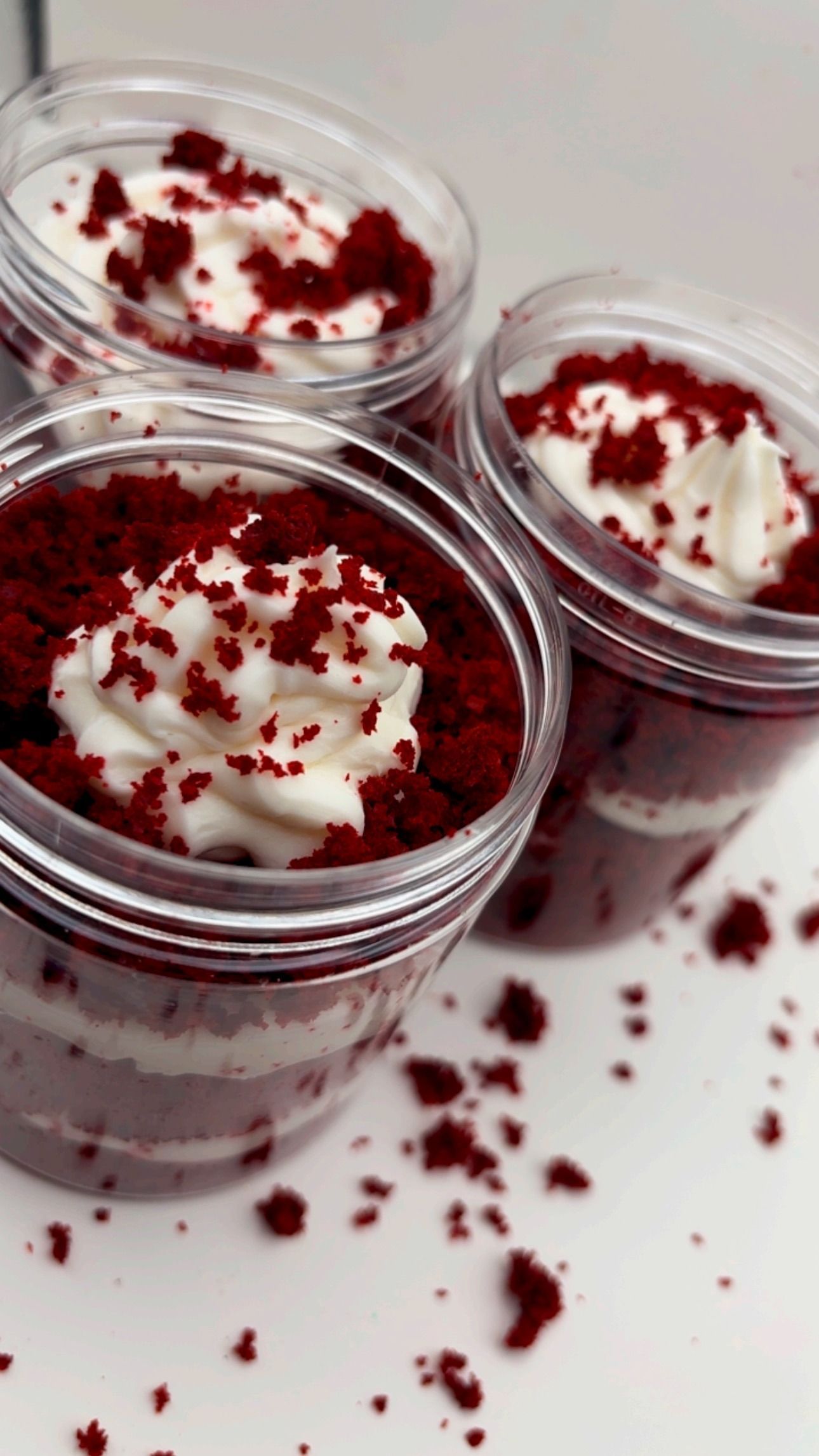 Three clear plastic jars filled with layered red velvet cake and white cream, topped with cake crumbs and a frosting swirl.