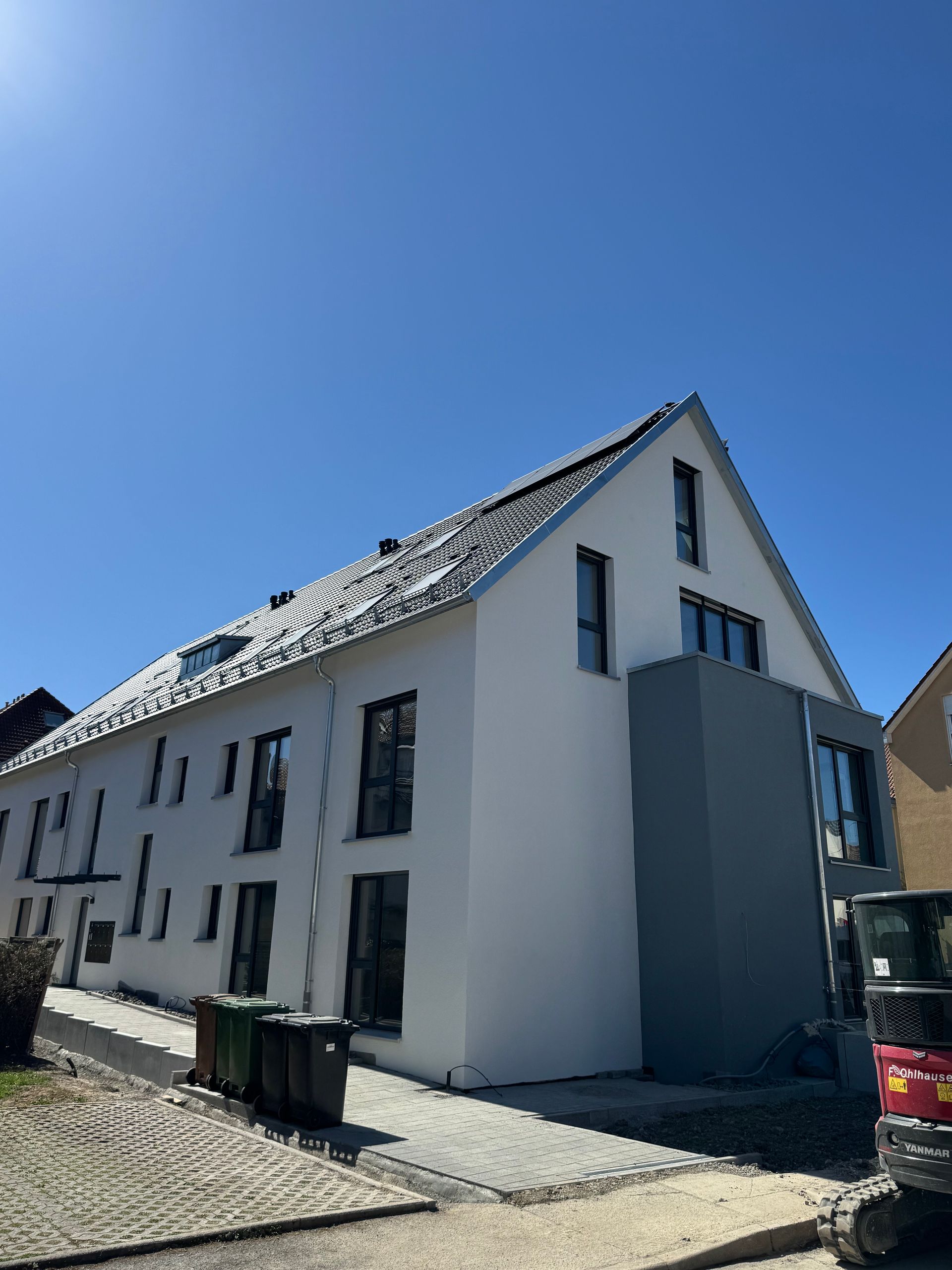 Weißes Gebäude mit dunklen Fenstern unter blauem Himmel. Grüne Mülltonnen davor, kleine Baumaschinen rechts.