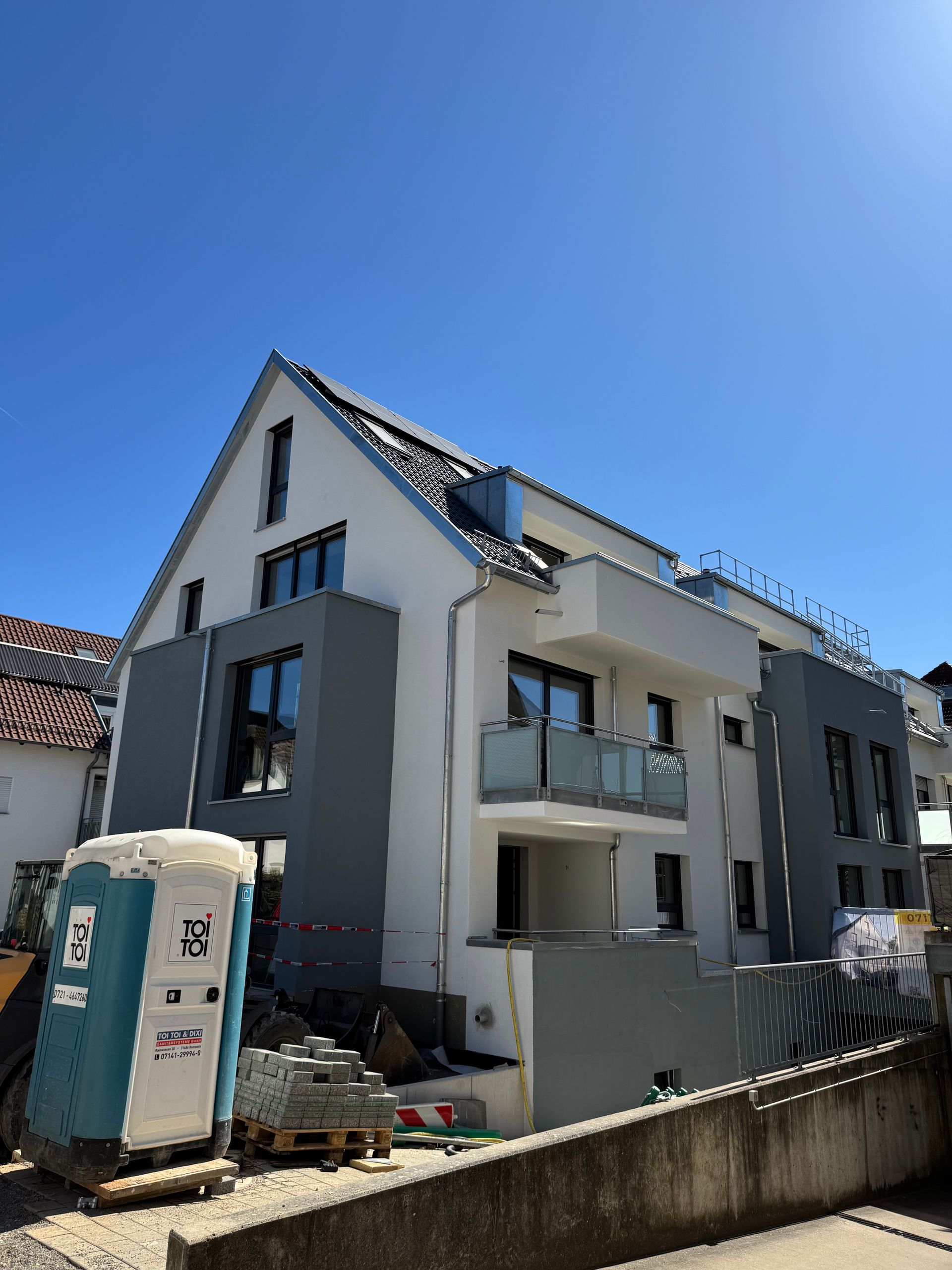 Gebäude im Bau, weiß-graue Fassade mit Balkon, davor eine blaue Mobiltoilette.