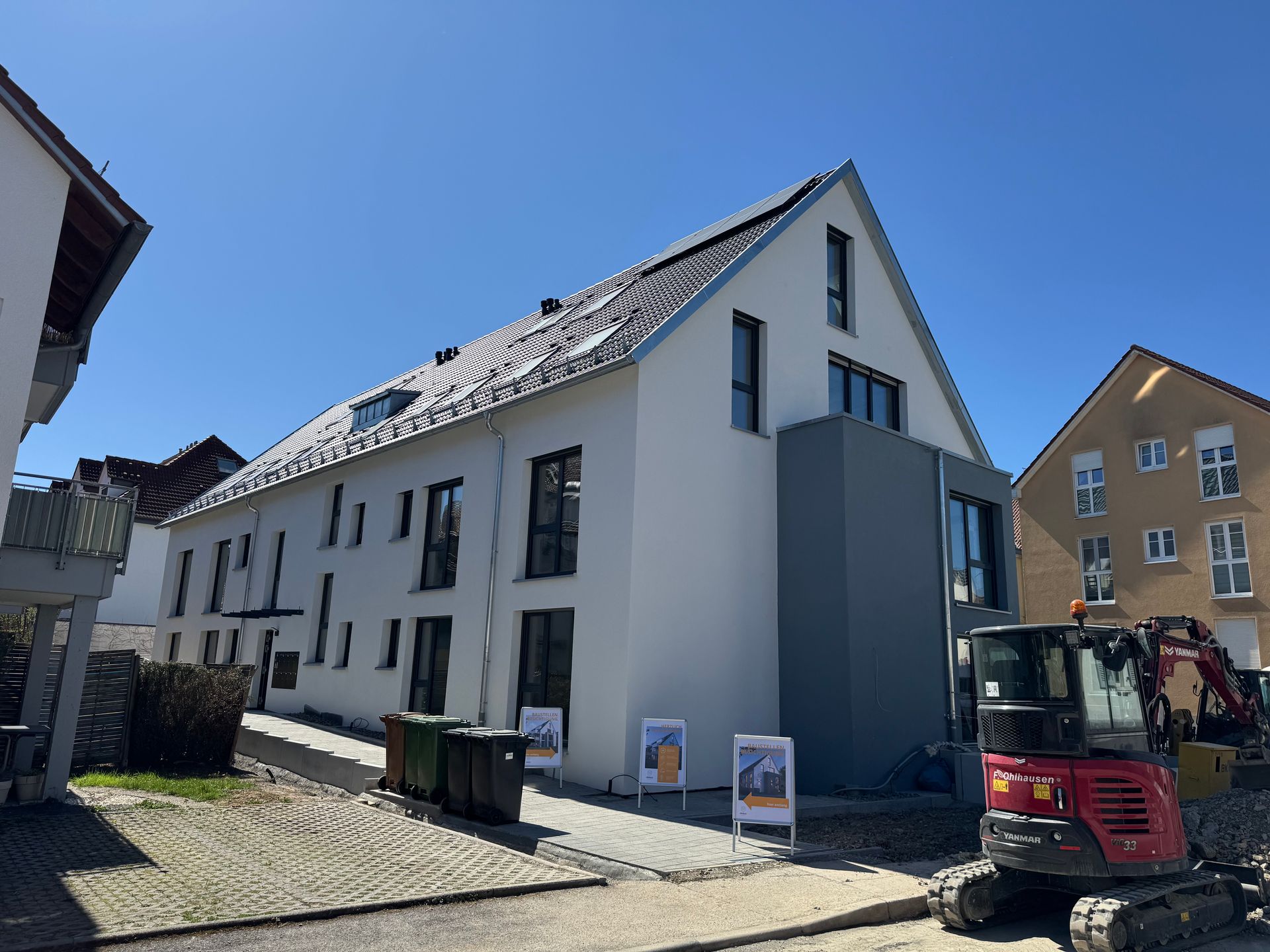 Mehrstöckiges weißes Gebäude mit dunklen Fenstern, grauen Akzenten, Solarmodulen, Baumaschinen; strahlend blauer Himmel.