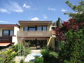 Ein großes Haus mit Balkon ist an einem sonnigen Tag von Bäumen und Büschen umgeben.