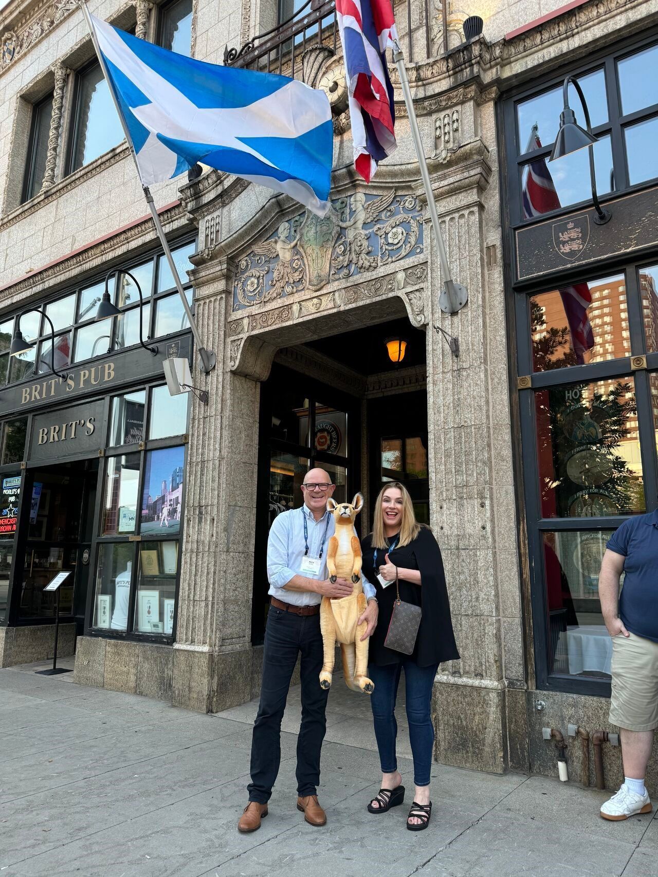 Two people pose with a light brown dog statue outside a building with flags.