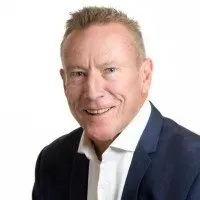 Man with short blonde hair smiles, wearing a navy blazer and white shirt.