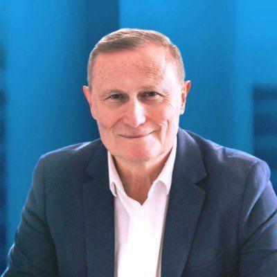 Man in navy blazer and white shirt, smiling. Blue background.