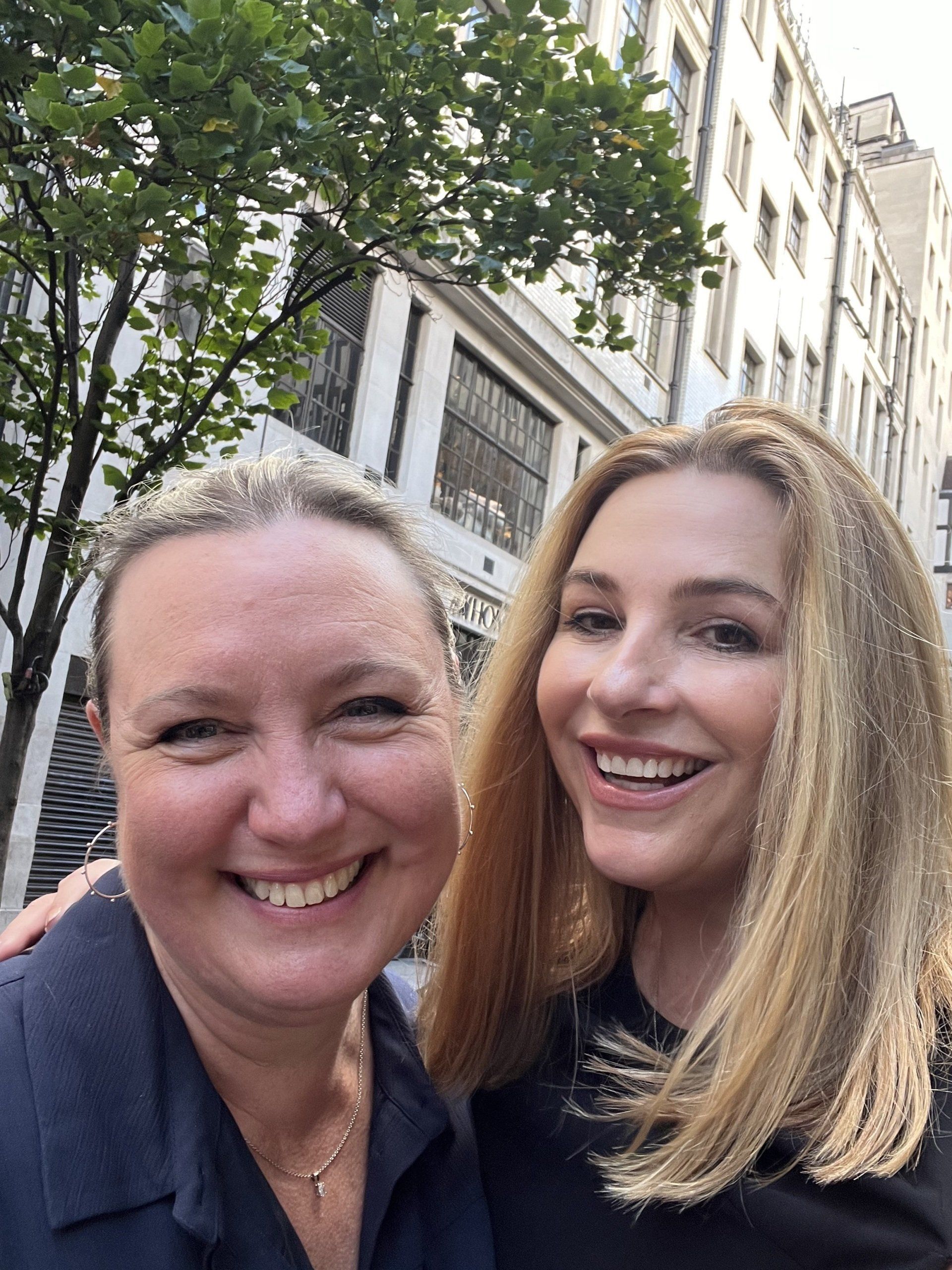 Two smiling women outdoors; one with blonde hair, the other with shorter, lighter hair. Buildings in the background.