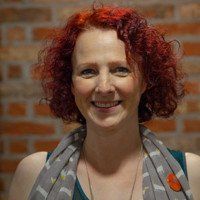 Woman with red curly hair smiles in front of a brick wall, wearing a gray scarf and green top.