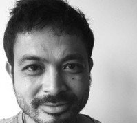 Close-up portrait of a man with dark hair and a beard, smiling slightly. Black and white.