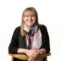 Woman with blonde hair smiling, wearing a scarf and black sweater, seated in a wooden chair.