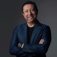 Man with arms crossed, smiling, wearing a blue blazer over a black shirt. Gray background.