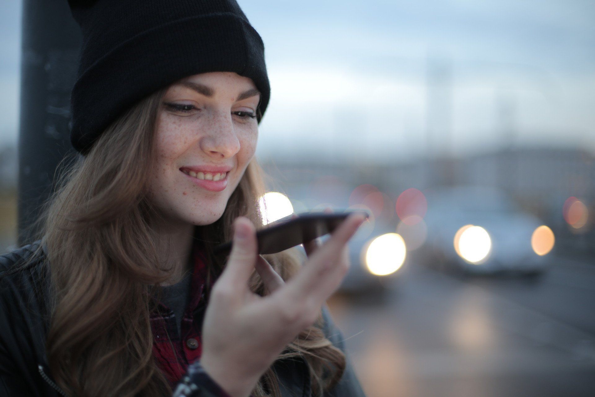 Young woman with freckles smiling, speaking into phone; city background with cars, lights.