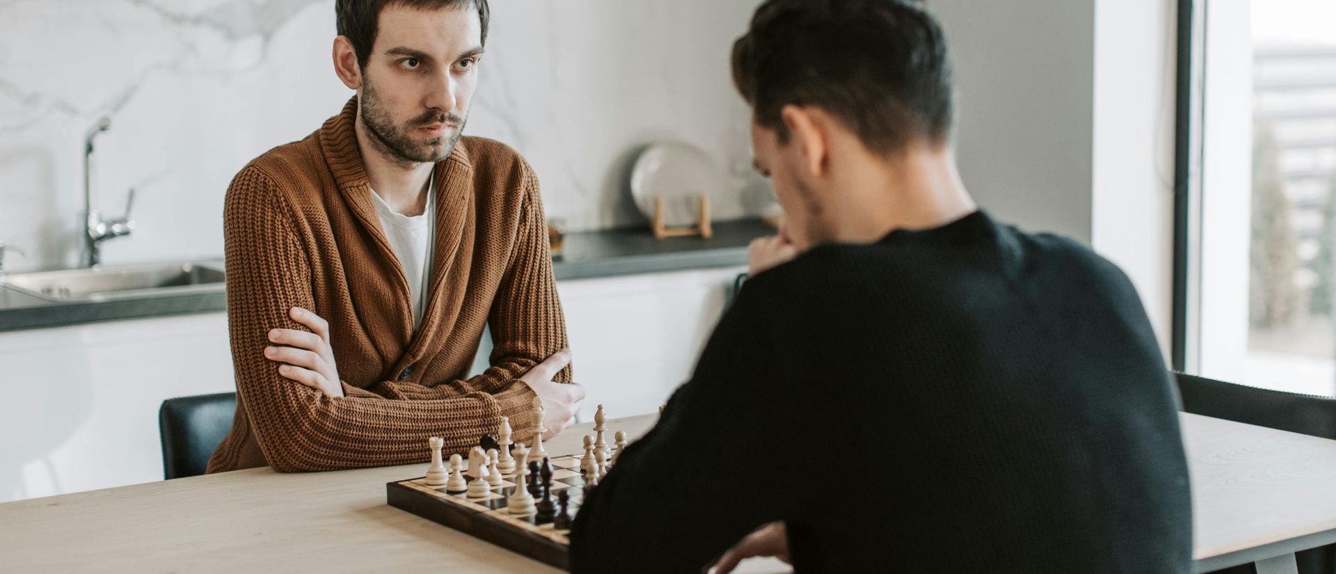 Two men playing chess at a table. One in a brown sweater, the other in black, set in a kitchen.