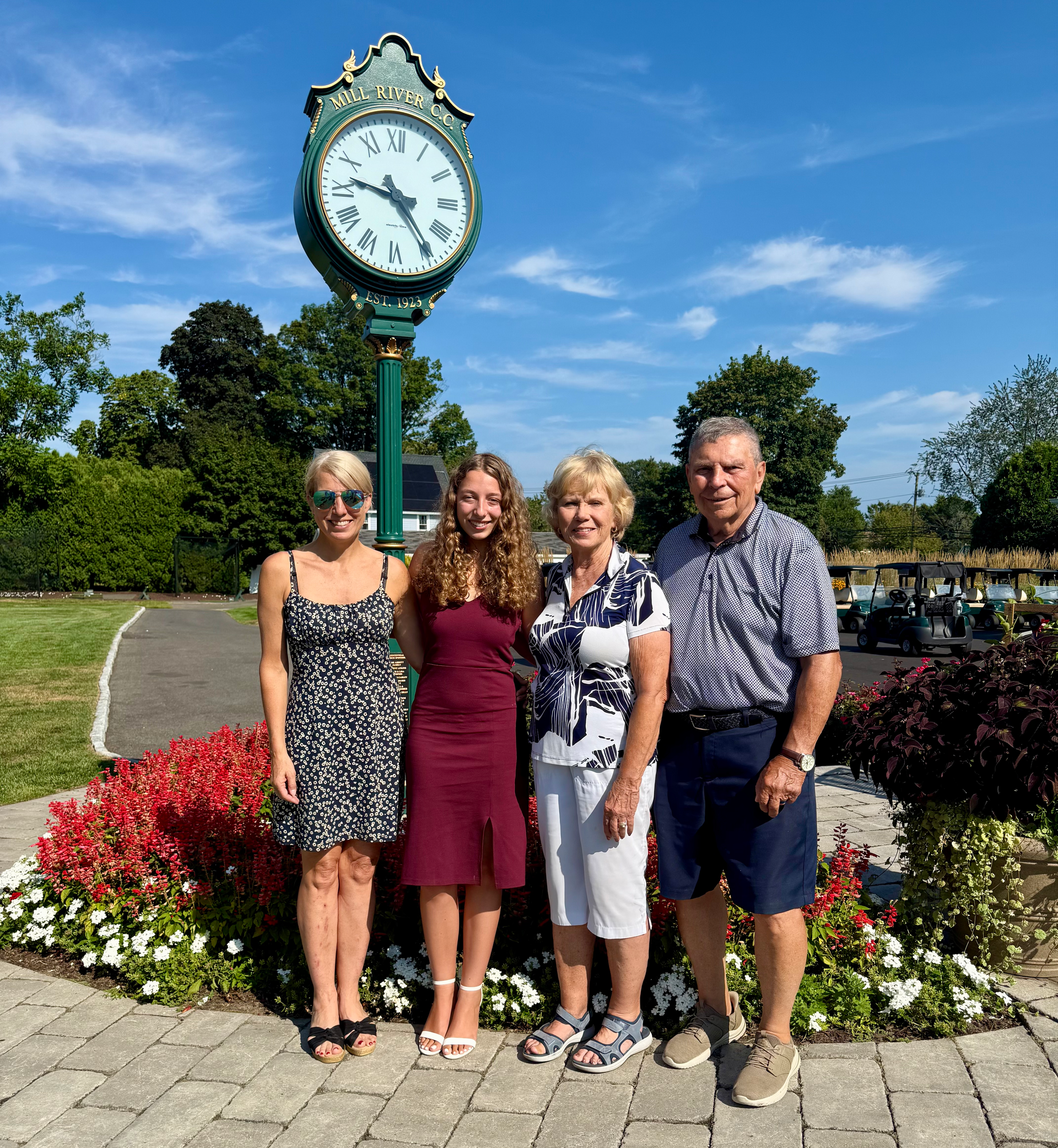 Four people pose in front of a large clock. Sunny day.