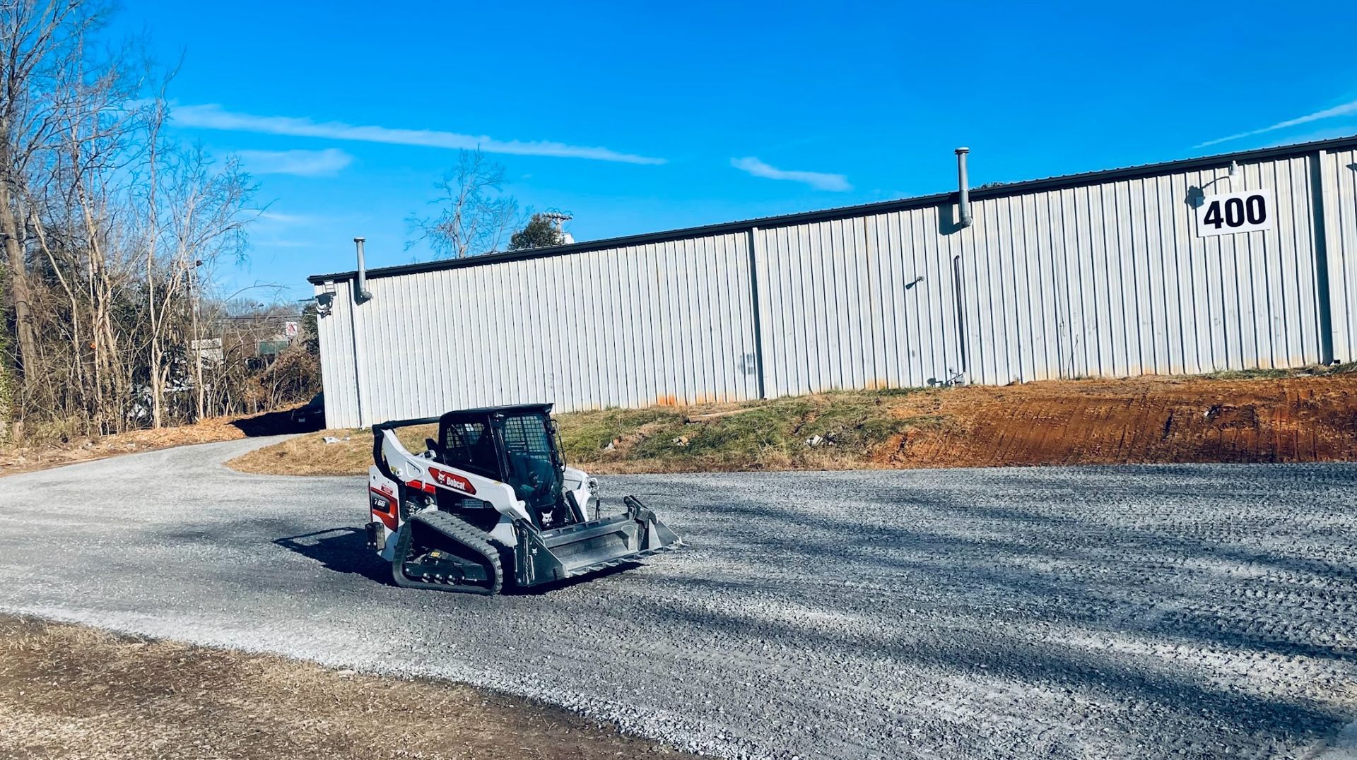 Skid steer loader on gravel road in front of a white warehouse with
