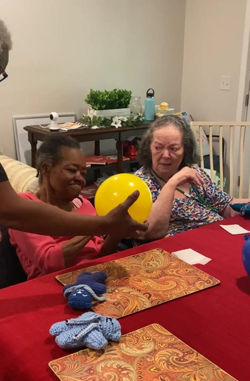 A person hands a yellow balloon to two seated individuals at a table.