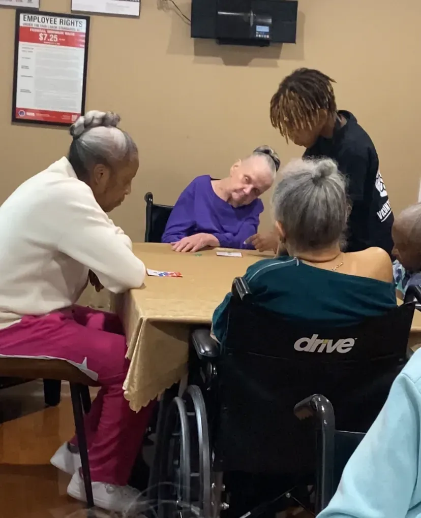 People playing a card game at a table. One person helps from behind.