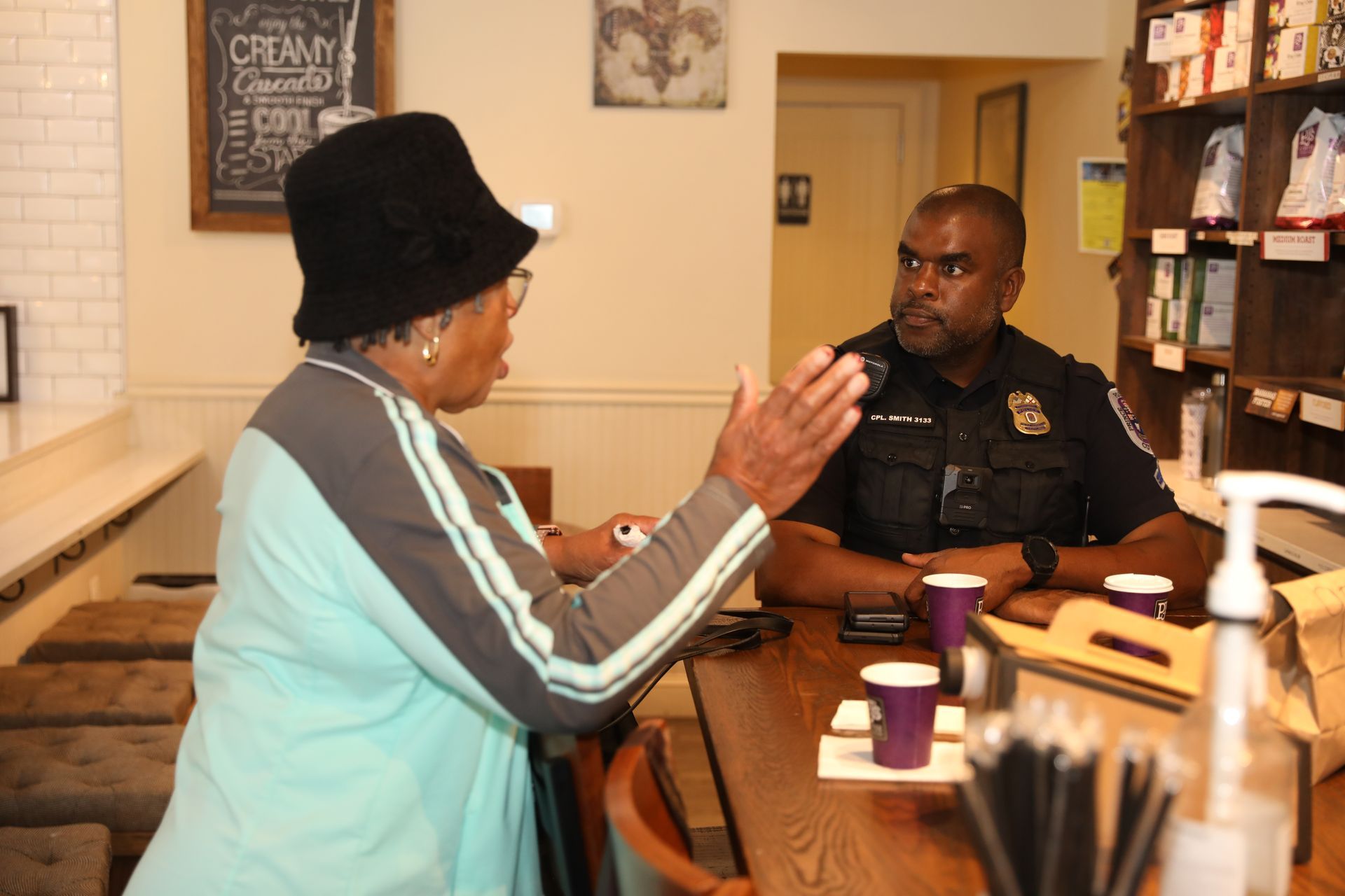 An individual in a black hat gestures while speaking to a uniformed police officer seated at a wooden counter.