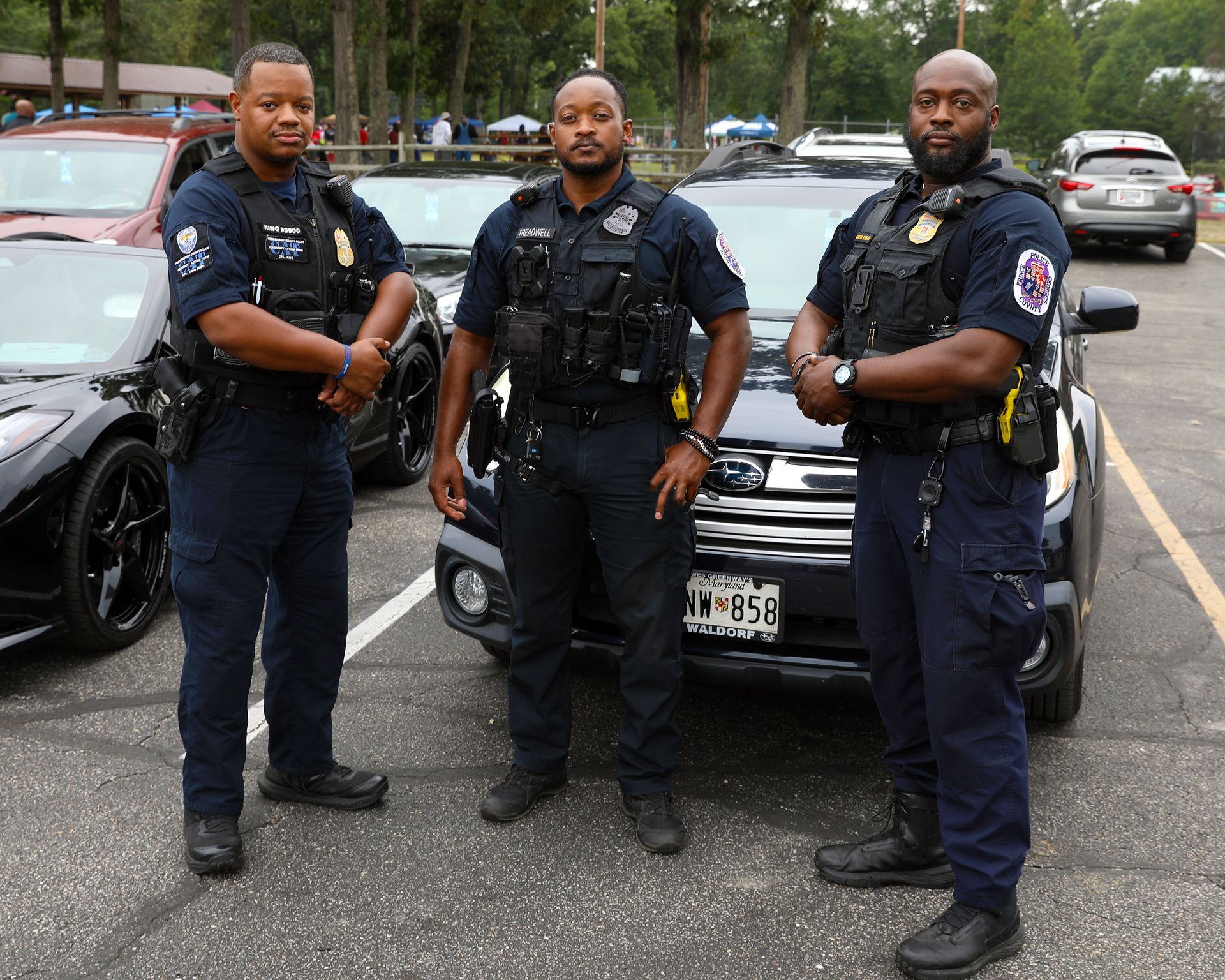 Three police officers in uniform stand in a parking lot, positioned in front of a dark Subaru patrol vehicle.