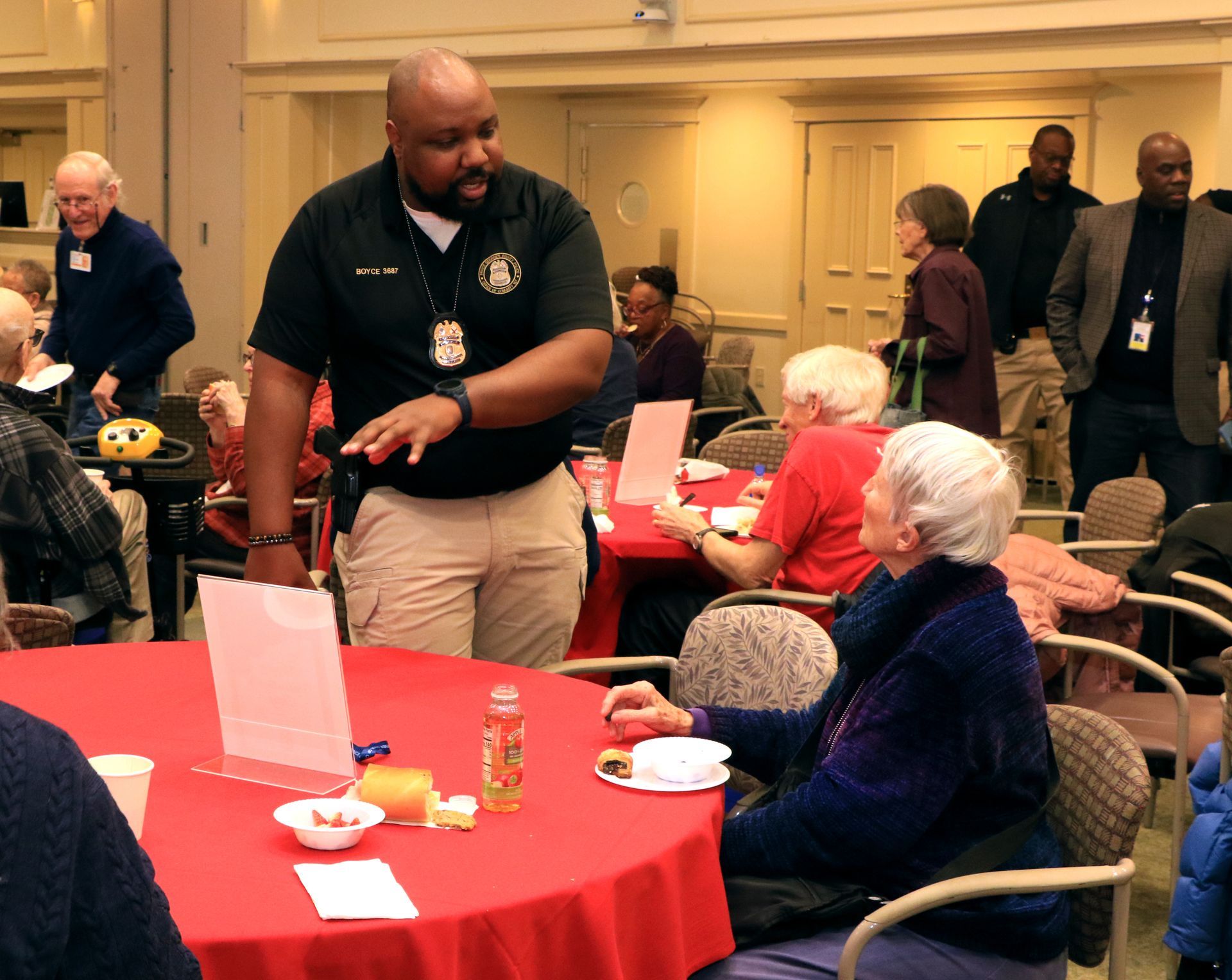 A speaker in a dark polo shirt gestures while presenting at a table in a room with attendees seated at red-covered tables.