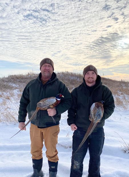 Two men are standing in the snow holding pheasants.