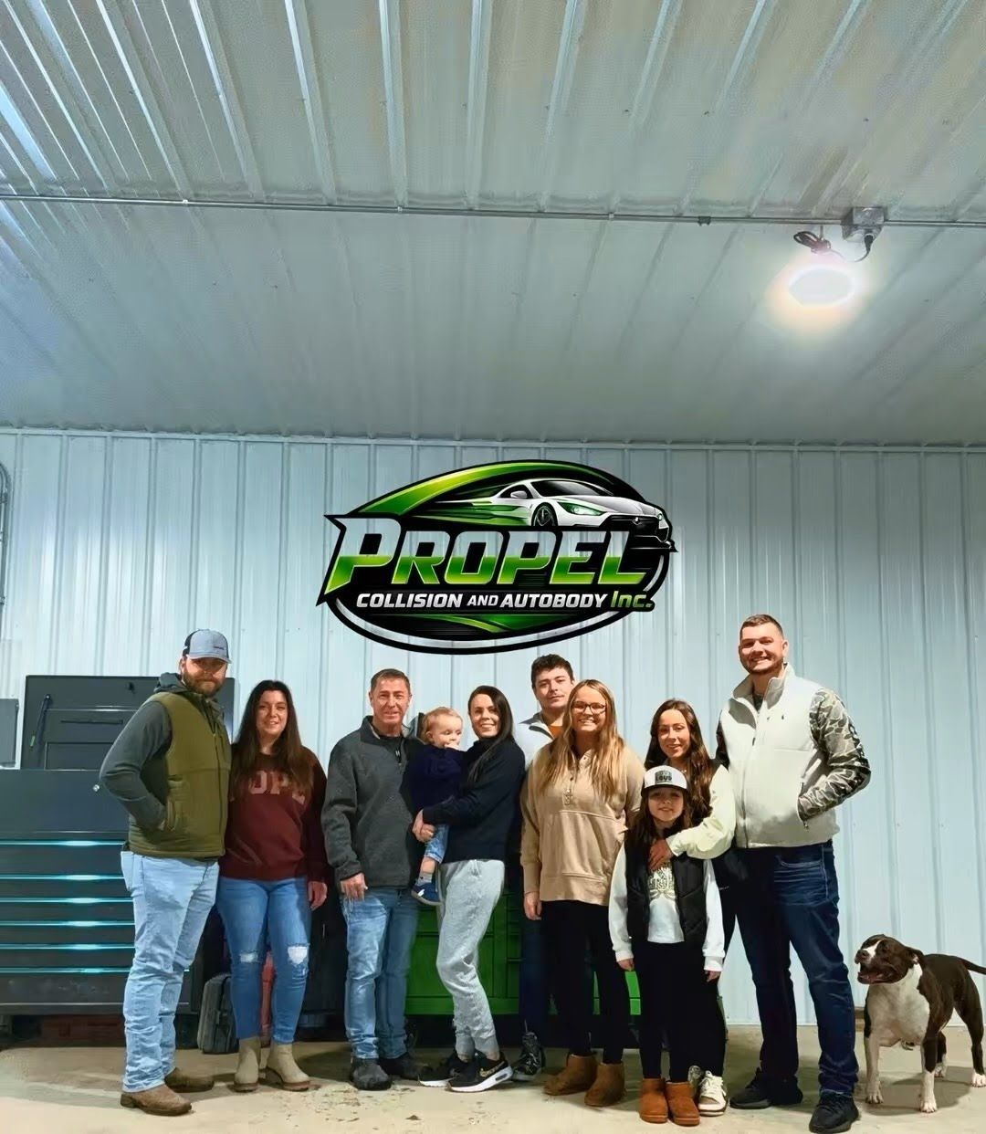 Group of people standing in front of a company logo. They are in a garage-like setting with a dog.
