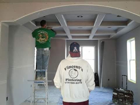 Two workers plastering an interior ceiling. One on ladder, other standing. Construction setting, gray walls.