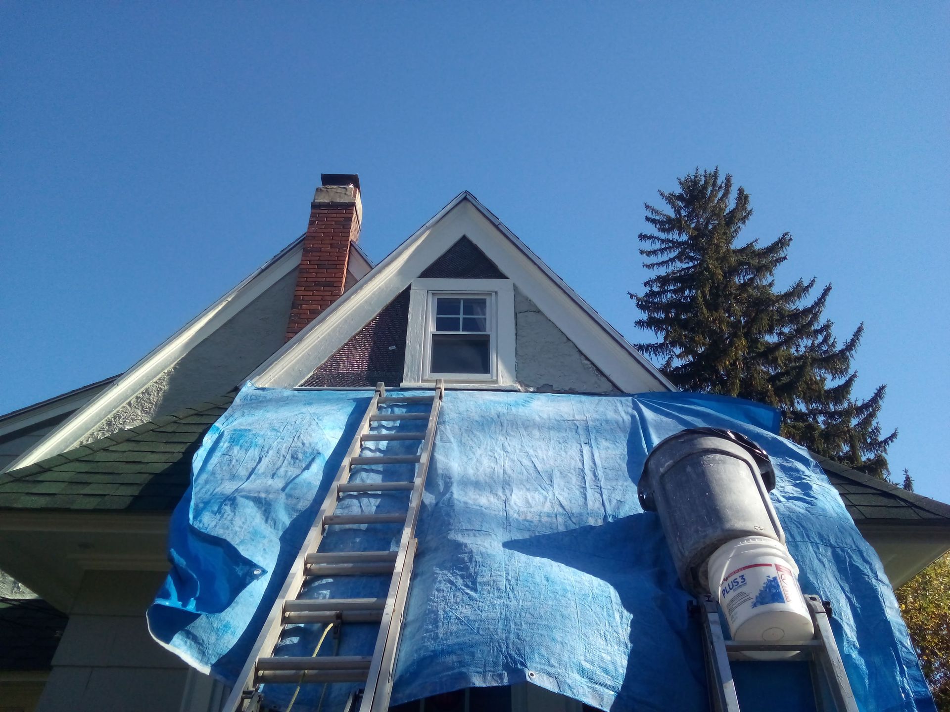 A house exterior with a ladder, tarp, and paint bucket against a blue sky.
