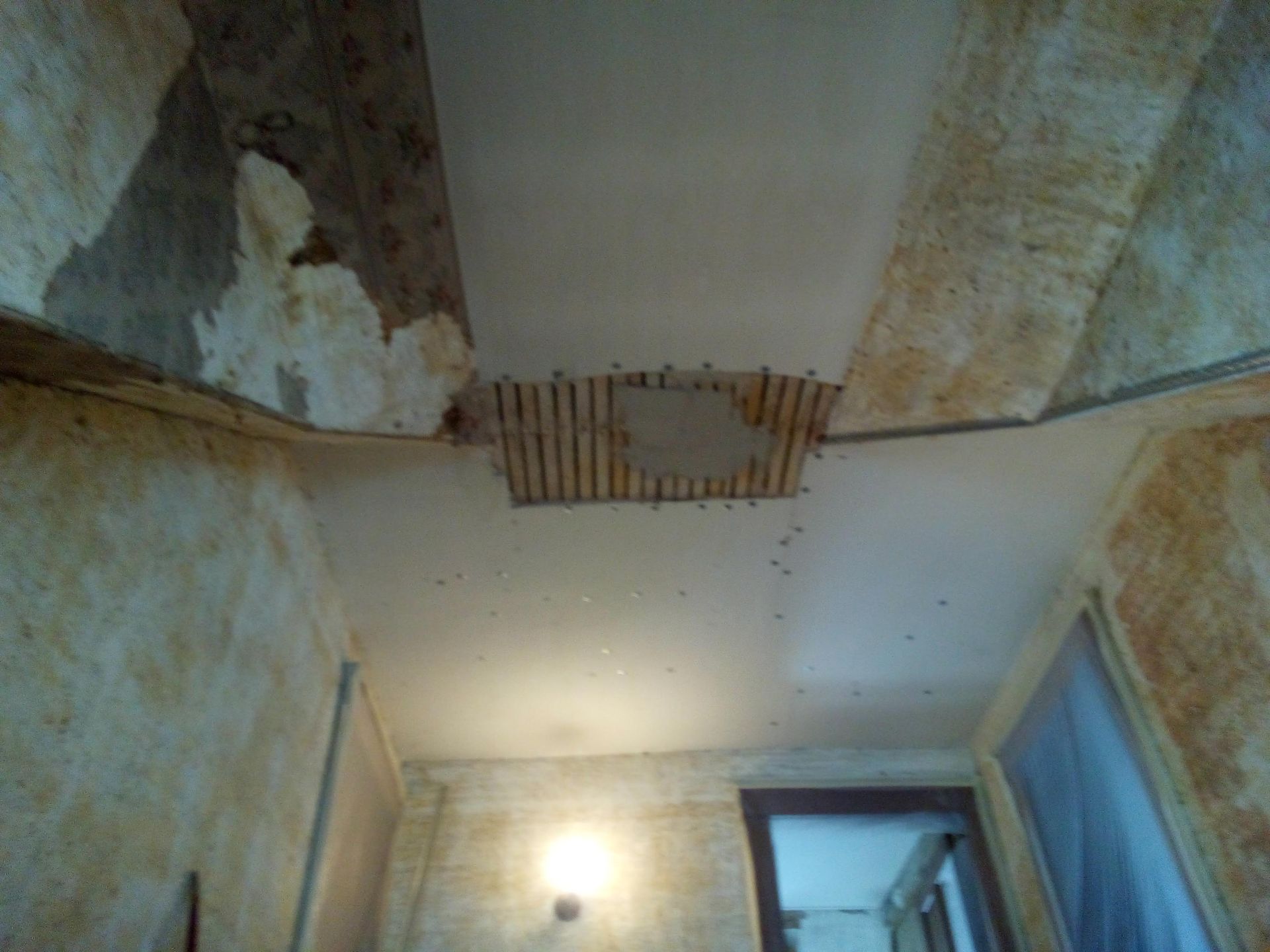 Ceiling damage, exposed wooden lath, and debris, in a room with peeling wallpaper and a light fixture.