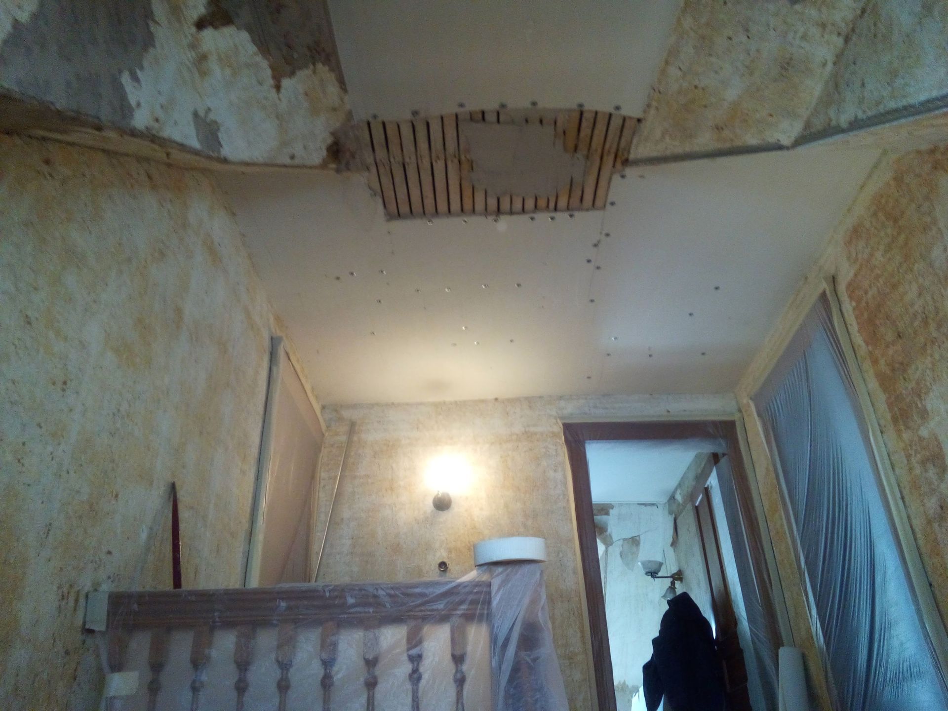Ceiling damage in a stairwell; hole in plasterboard reveals wooden slats; walls and staircase covered in protective sheeting.