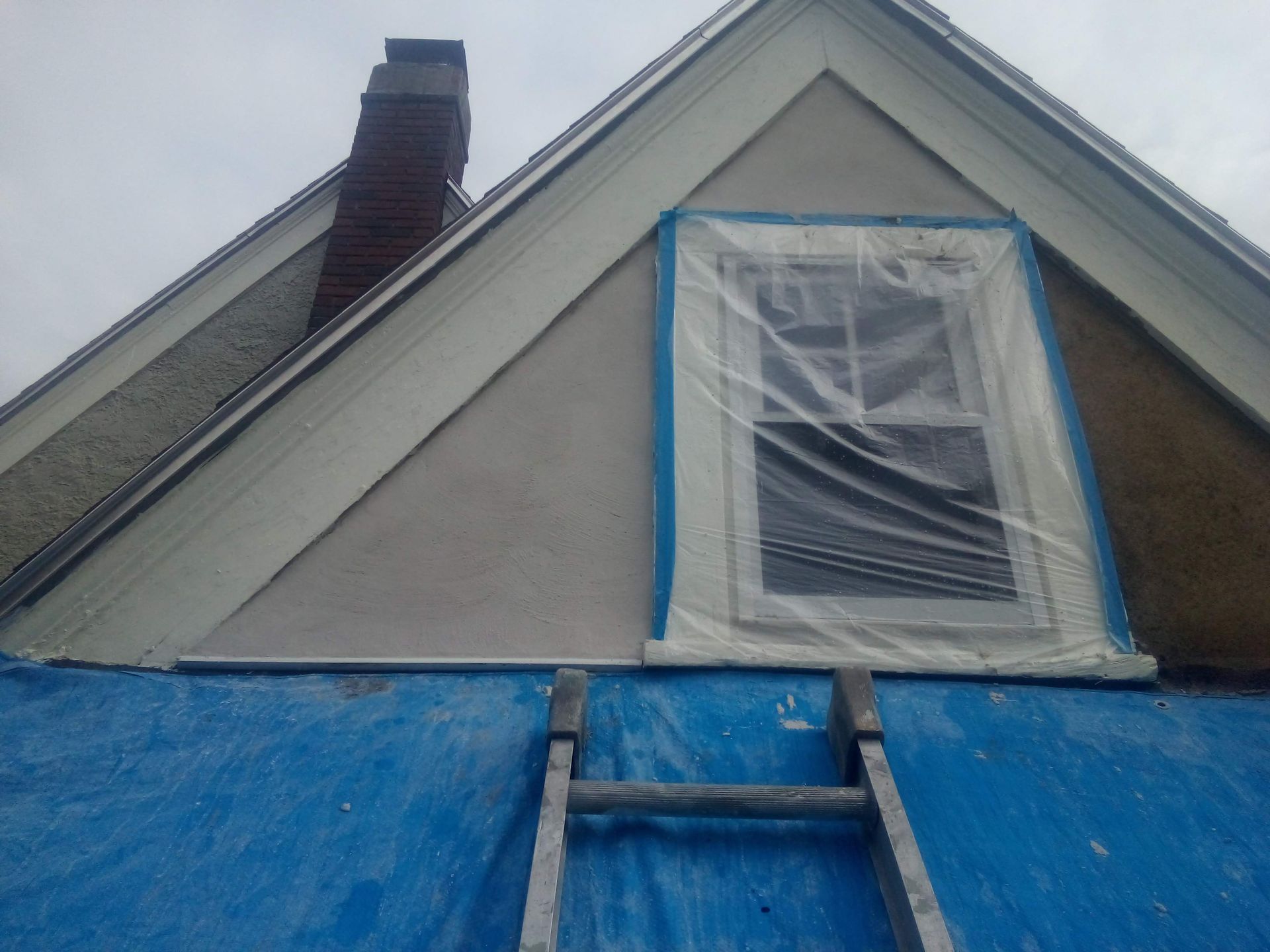 Exterior house with window taped and ladder against blue tarp; stucco wall.