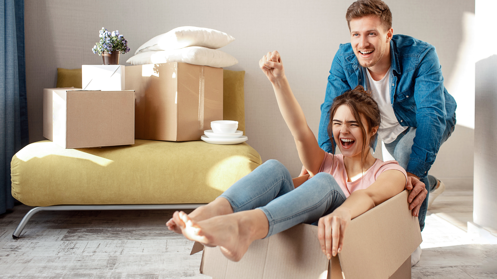 A couple having fun during their move, with the woman sitting inside a cardboard box and the man pushing her, both smiling and enjoying the moment in their new home.