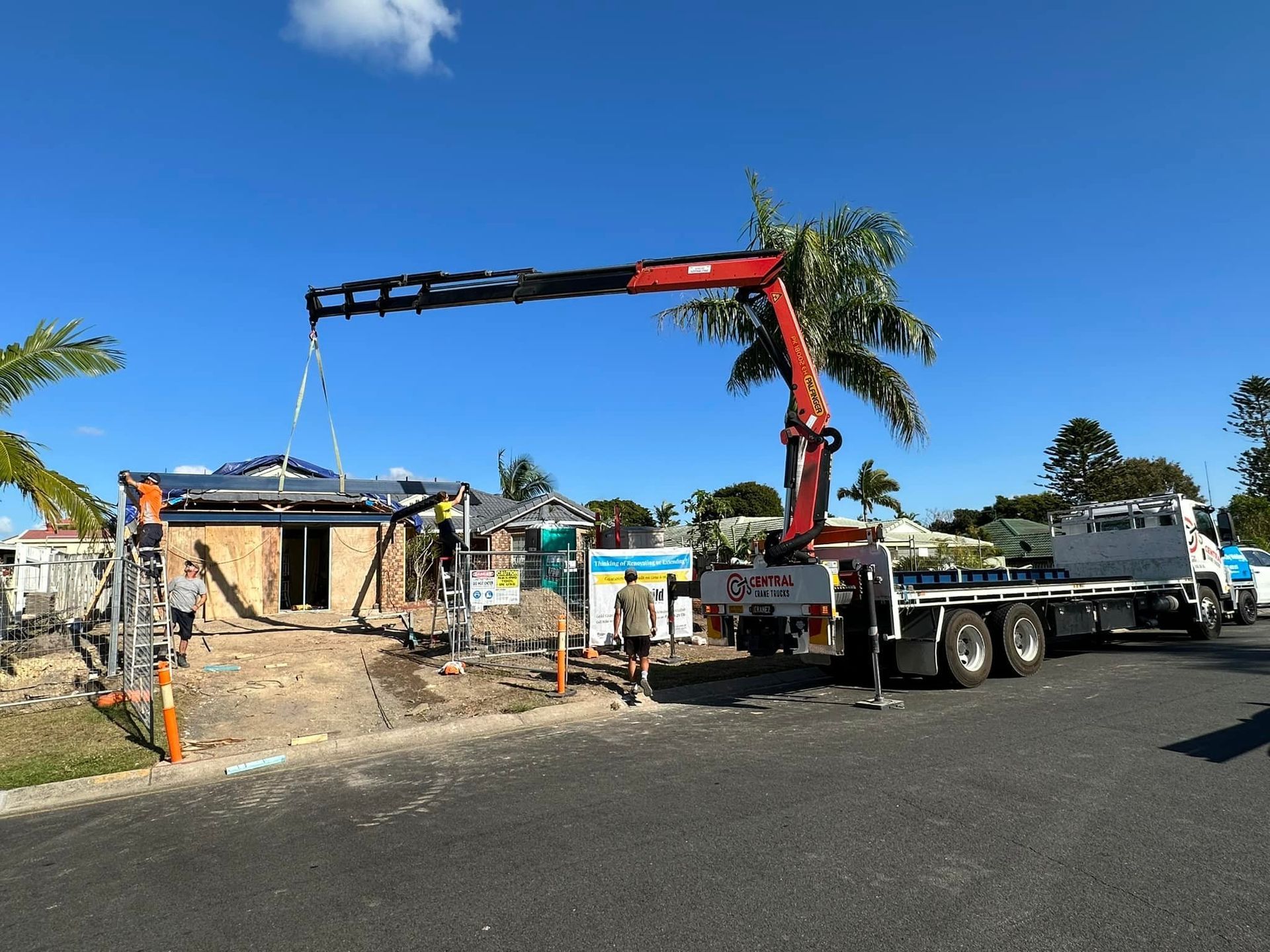 Crane truck lifting roof trusses onto a house under construction with workers on site.