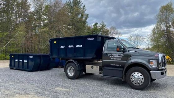 A dump truck is parked in a gravel lot next to a dumpster.