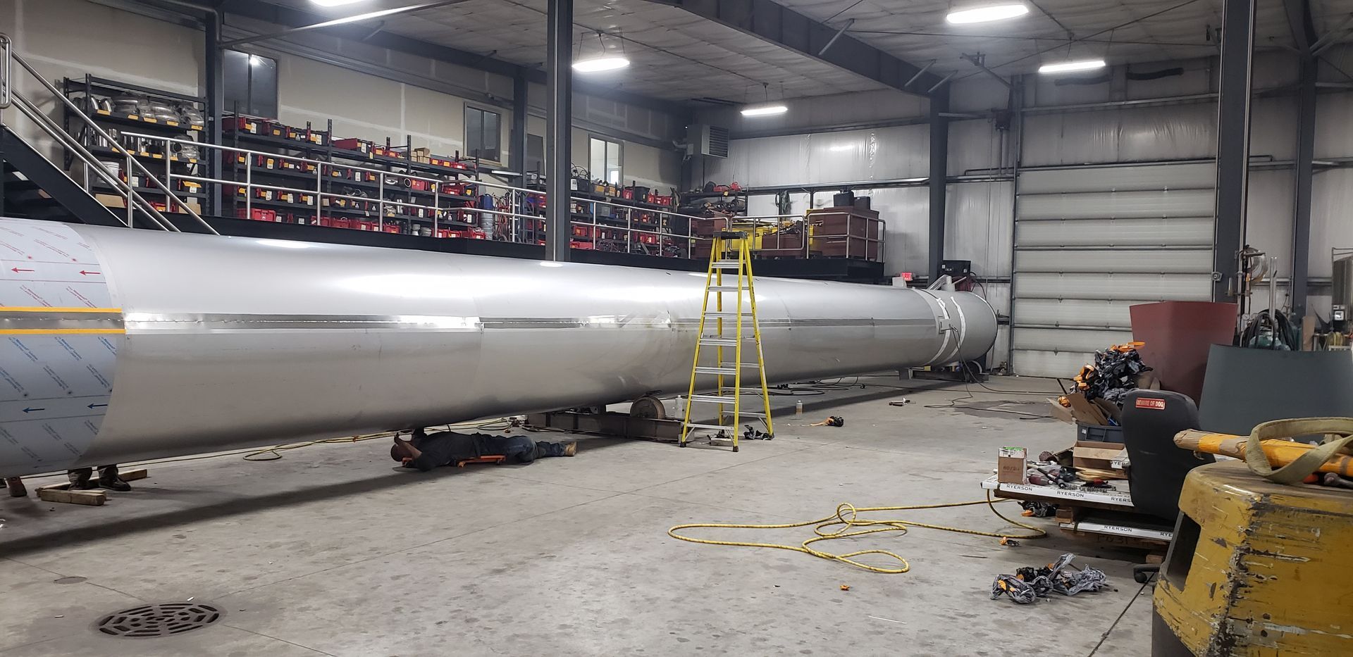 Inside a large industrial workshop, a long metal cylinder is being worked on by a ladder.