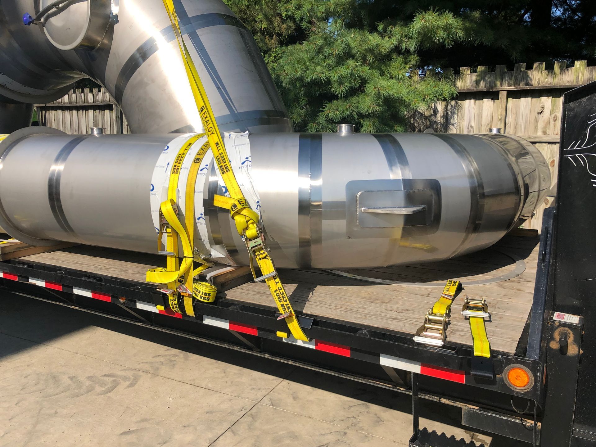 Large stainless steel pipe segment secured on a flatbed trailer with yellow straps.