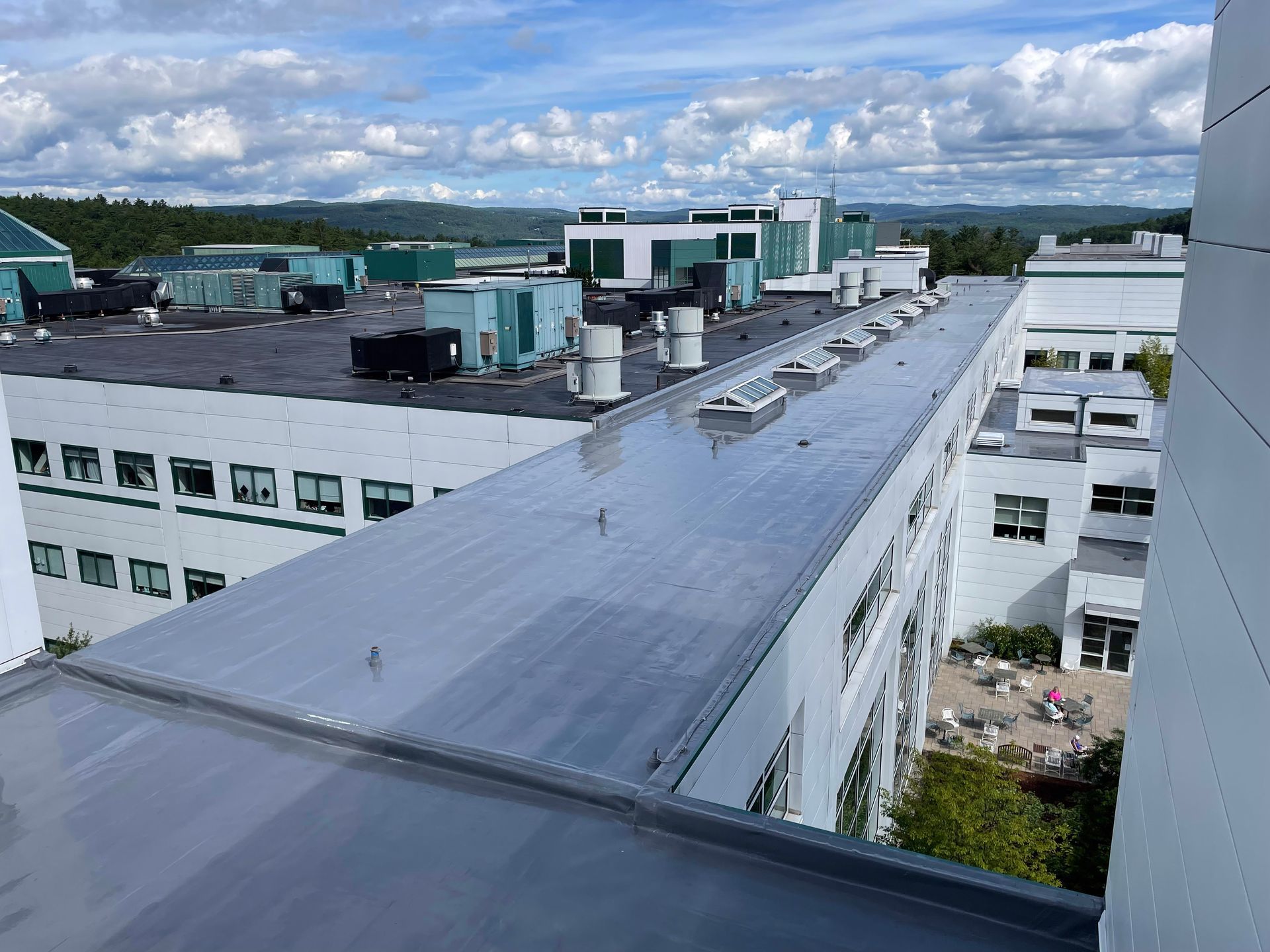 Overhead view of a building with a flat, reflective roof and multiple ventilation units. Cloudy sky in the background.