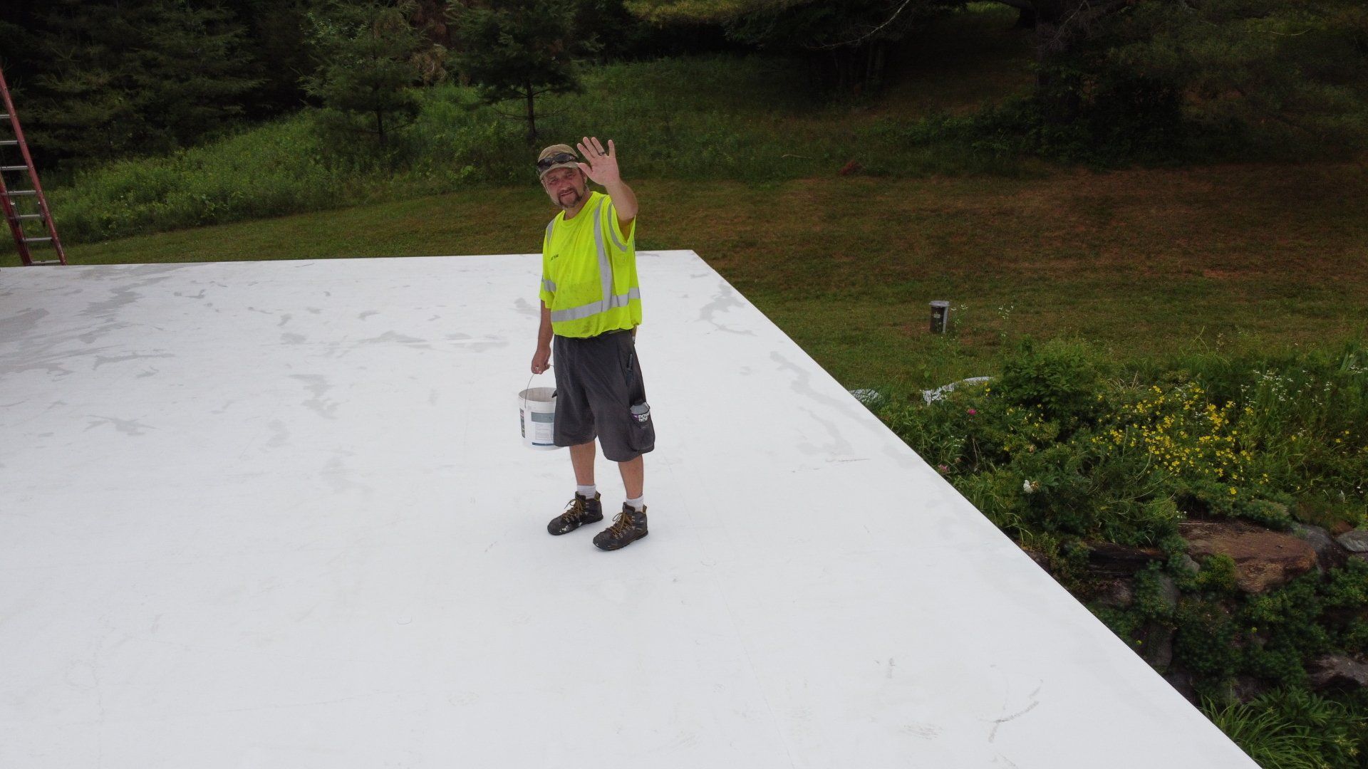 A man is standing on top of a white roof holding a bucket of paint.