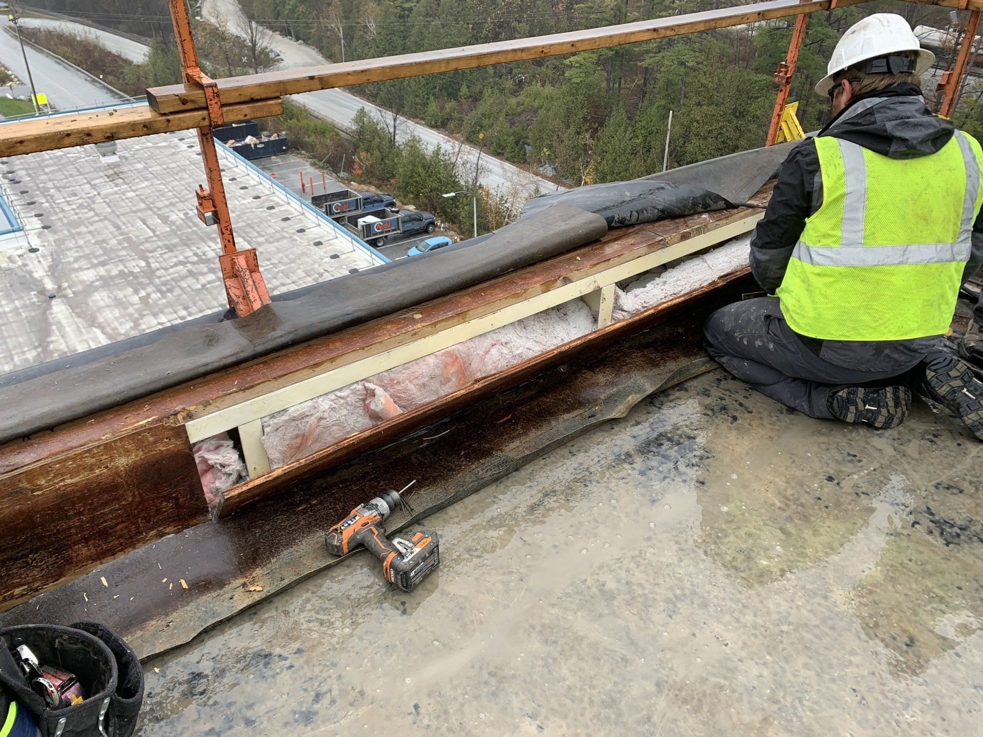 A construction worker is kneeling on a roof with a drill.