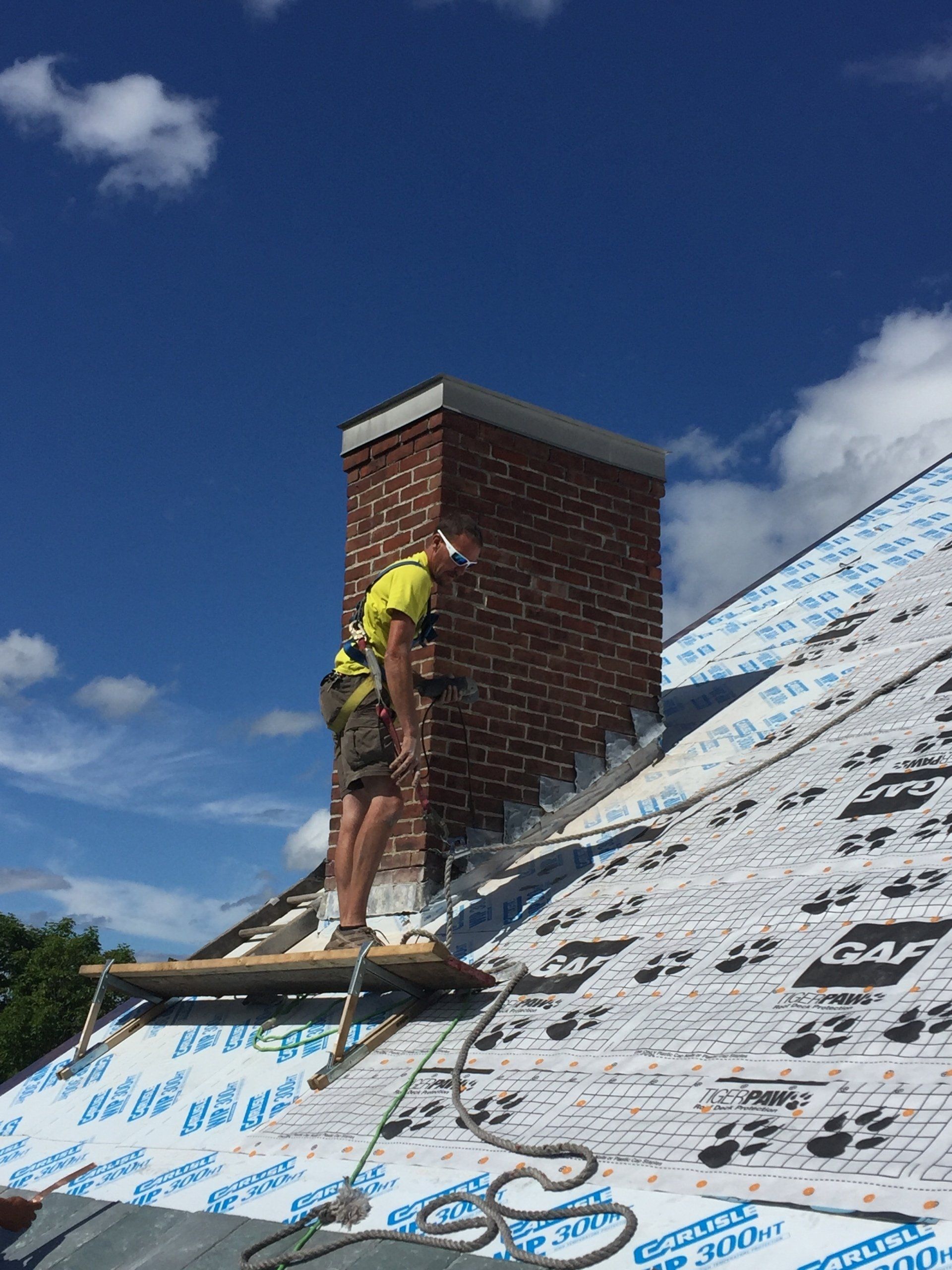A man is standing on top of a roof with a brick chimney.