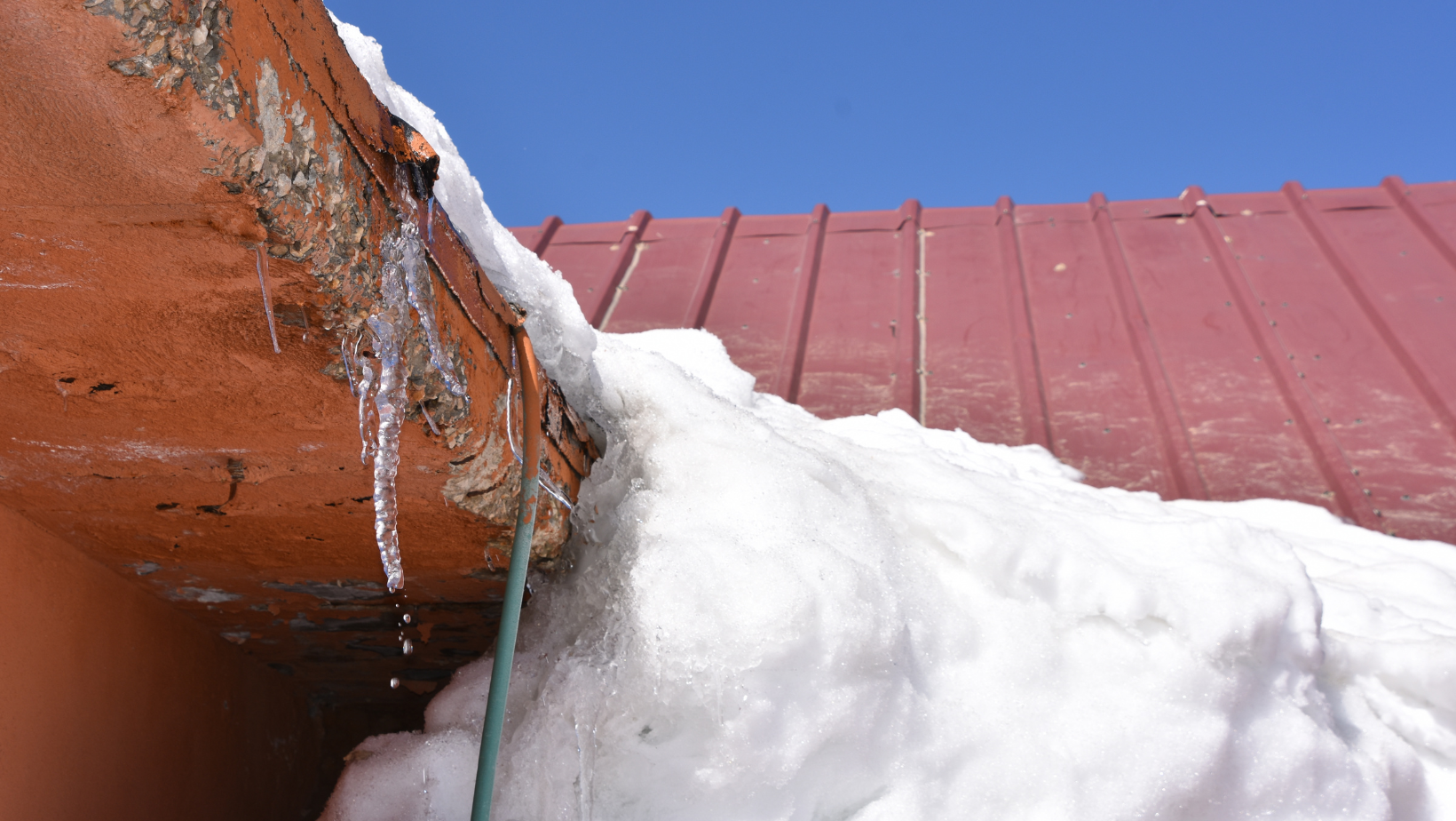 Melting snow and an icicle hang from a red metal roof under a bright blue sky.