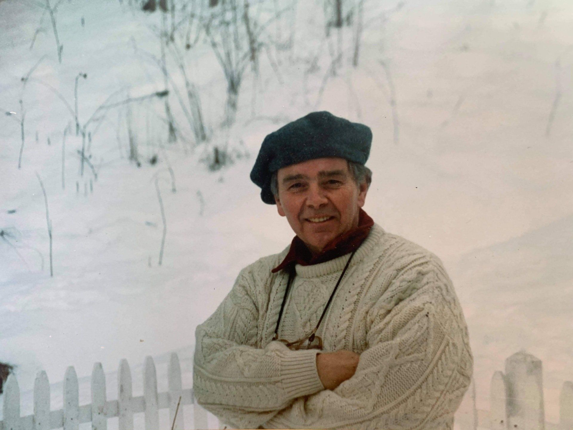 A man wearing a sweater and beret is standing in the snow with his arms crossed.