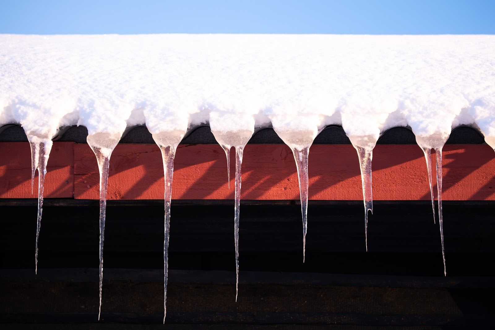 Snow-covered roof with icicles hanging from the edge, red siding below, against a clear blue sky.