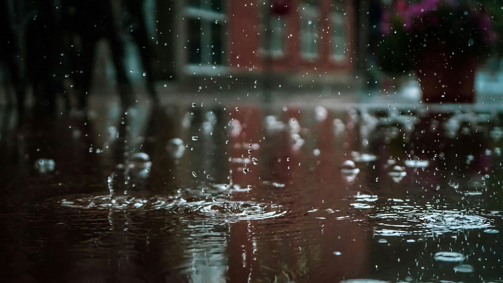 Raindrops splash into deep, shimmering puddles on a wet pavement with a blurred red building in the background.