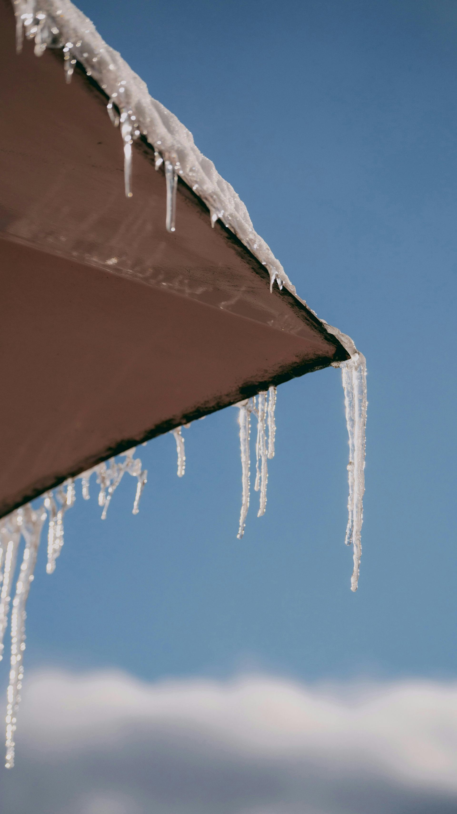 Icicles hanging from the dark brown edge of an umbrella against a clear blue sky.