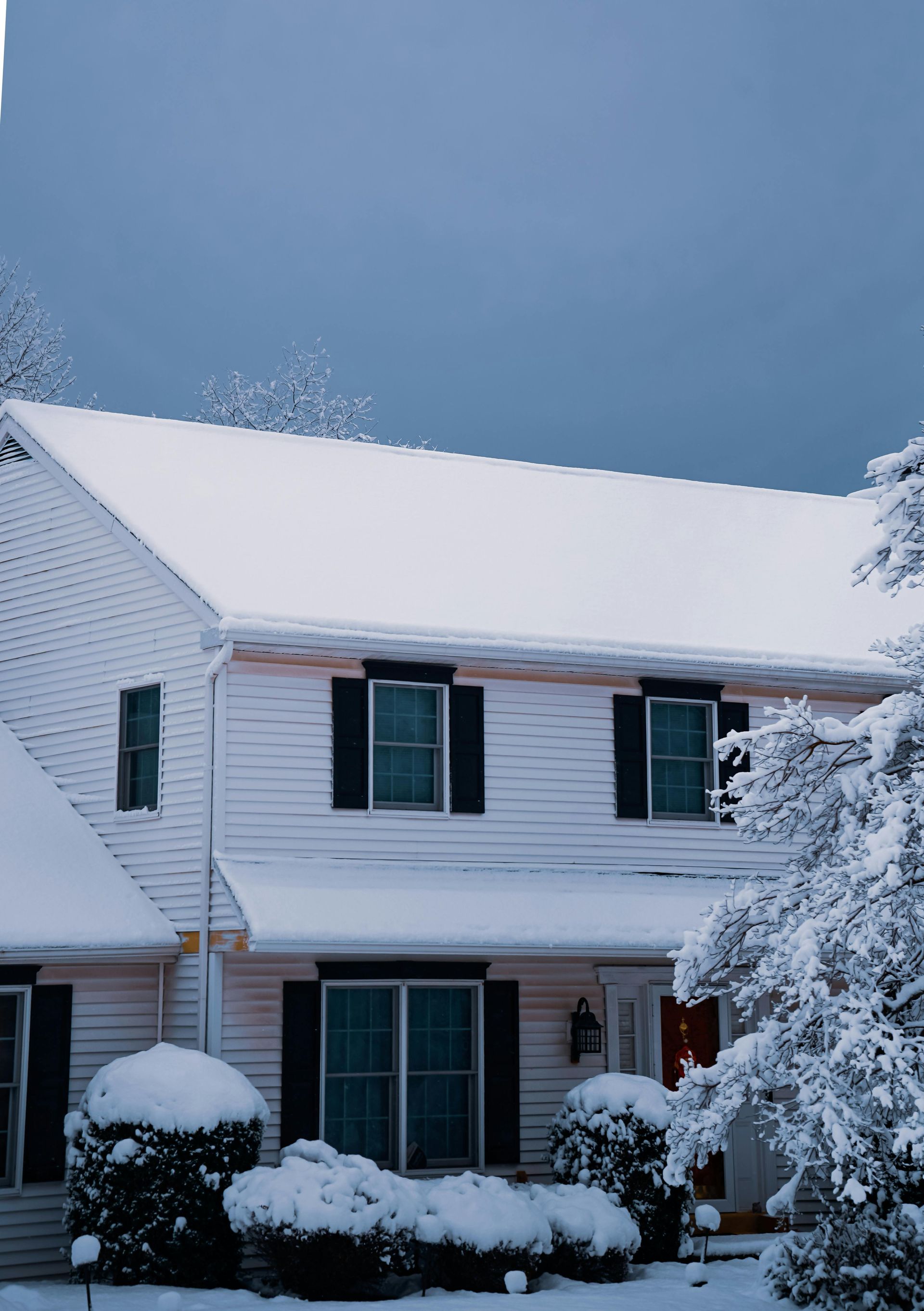 Two-story house covered in snow, with black shutters and a red door, during a winter day.