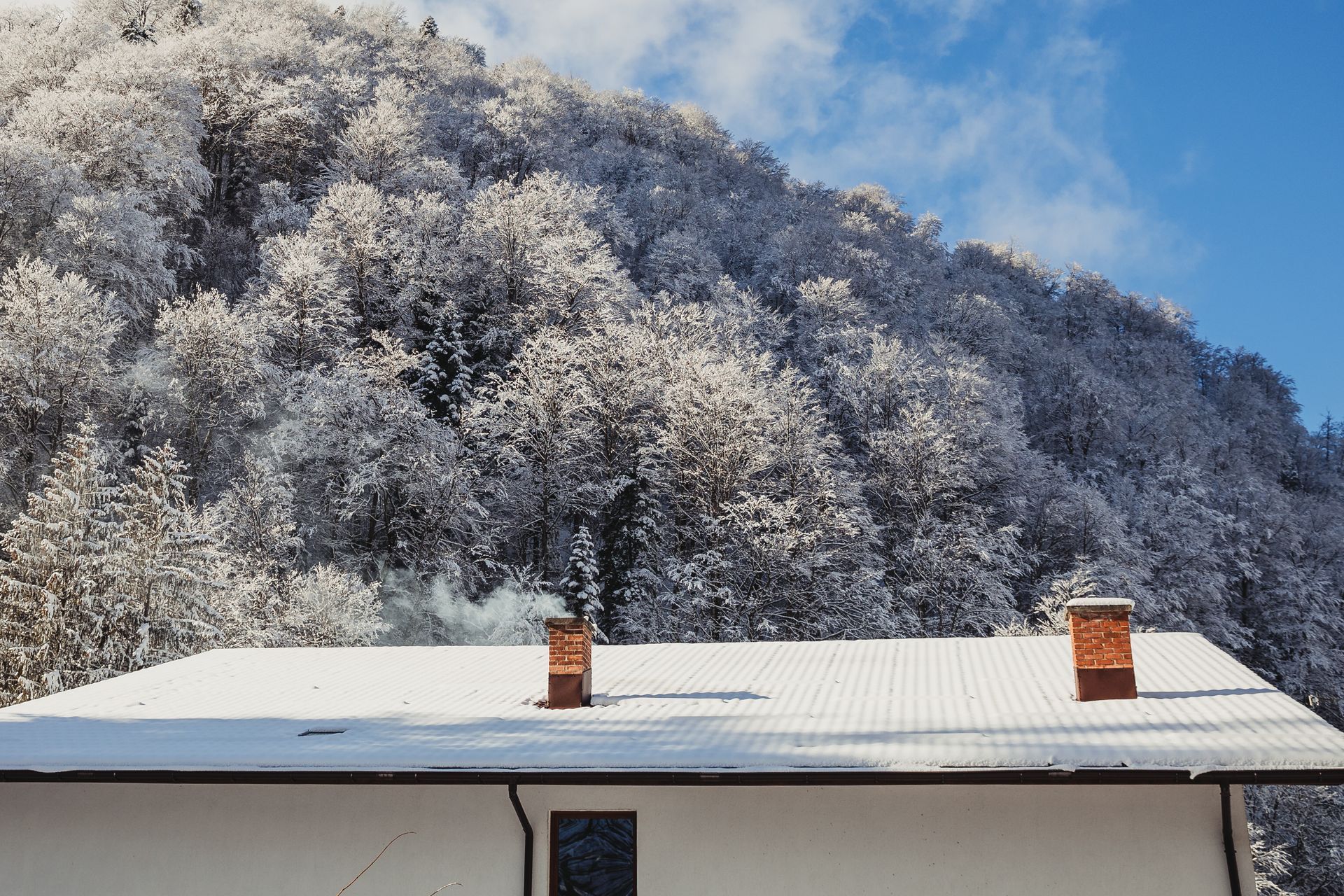 Snow-covered roof with smoking chimneys in front of a snowy forest on a sunny day.