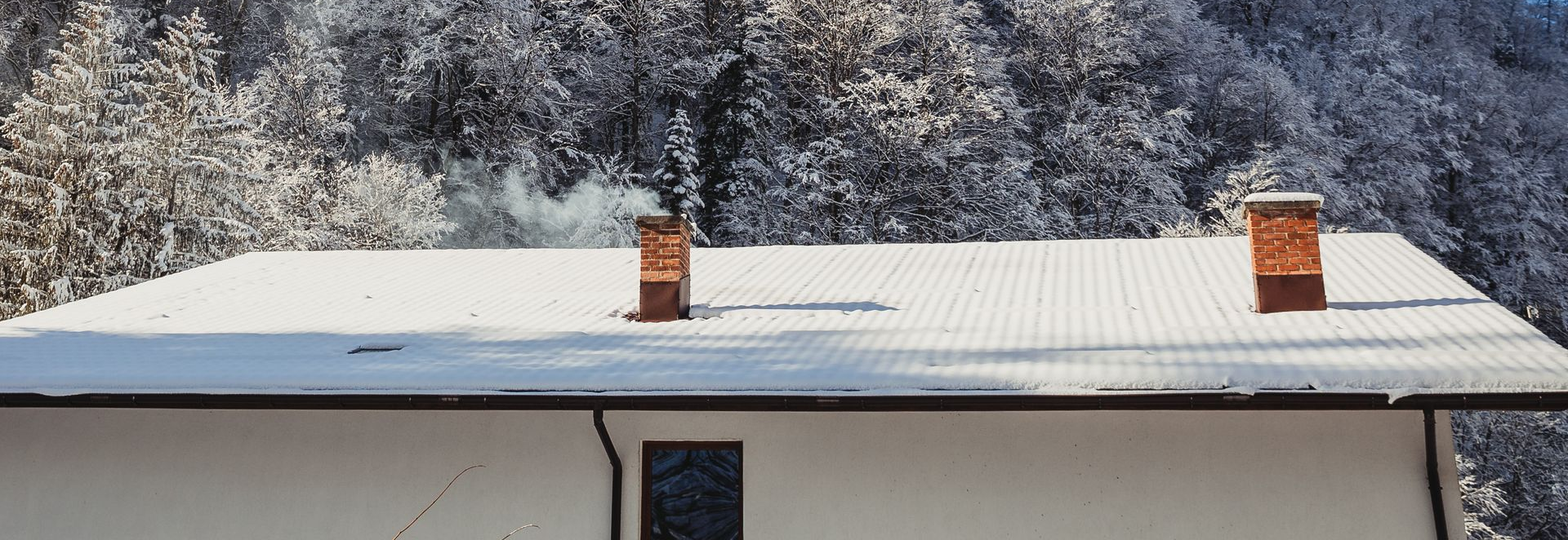 A snow-covered roof with two chimneys, smoke rising against a backdrop of snowy trees and a bright blue sky.