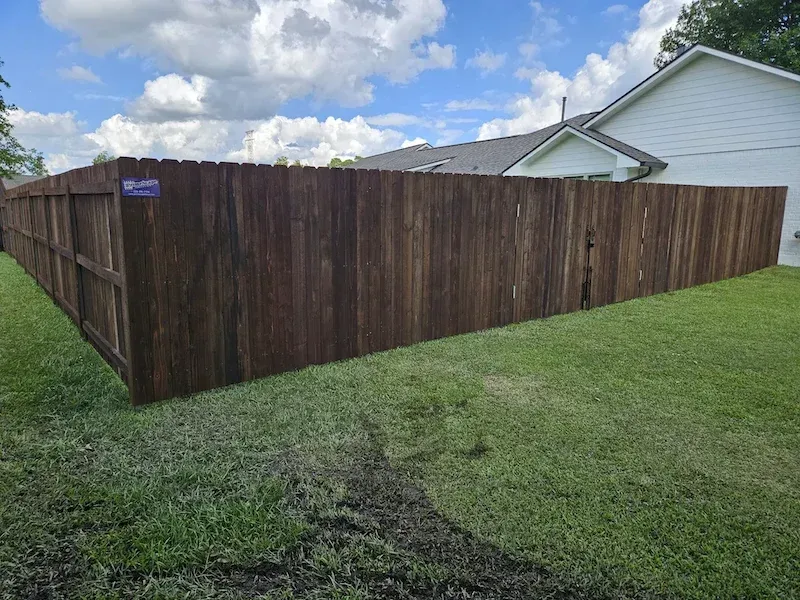 Dark wooden privacy fence around a grassy yard beside a white house under a cloudy sky