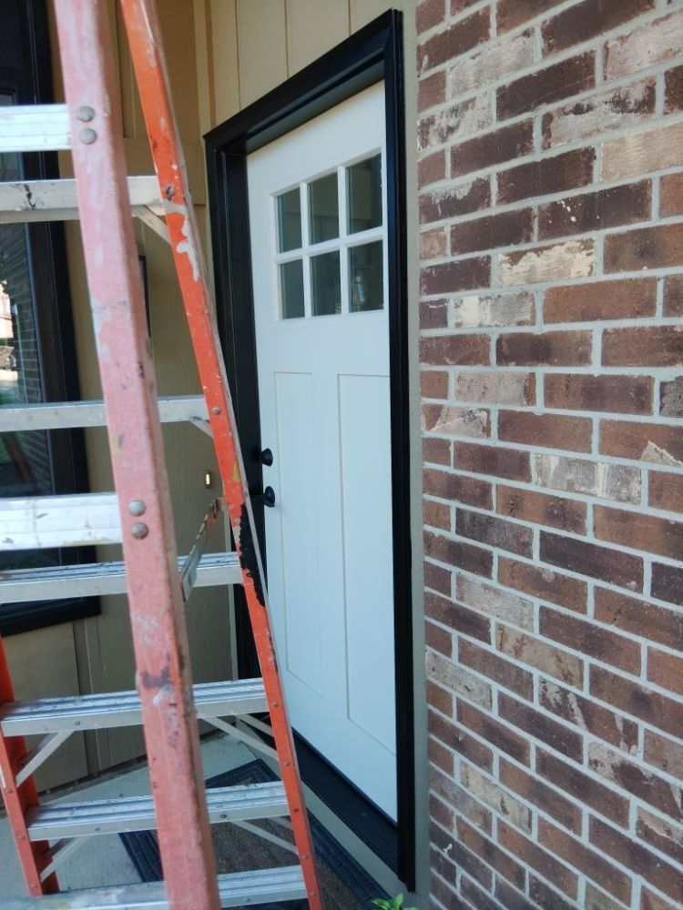 A white front door with a window and black trim, a ladder leans against the door frame, brick wall.